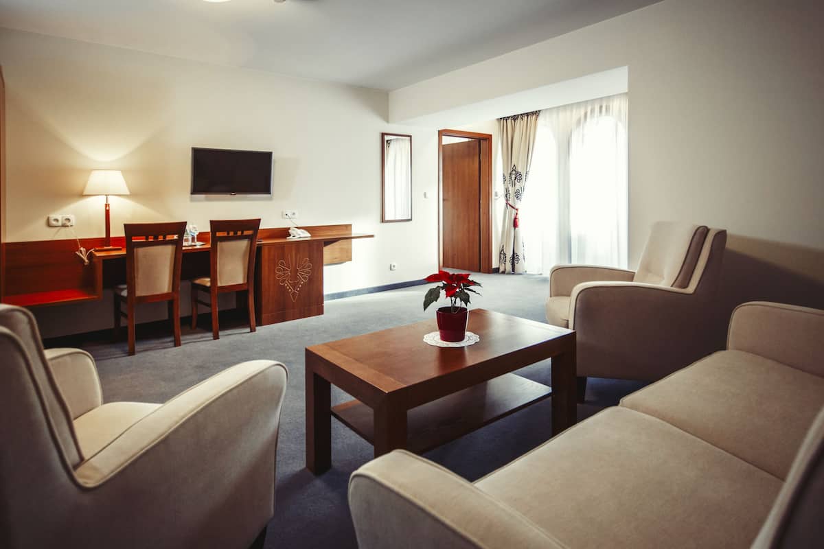 Quarto de hotel com área de estar, mesa de jantar com cadeiras, TV fixada na parede e uma janela grande com cortinas.