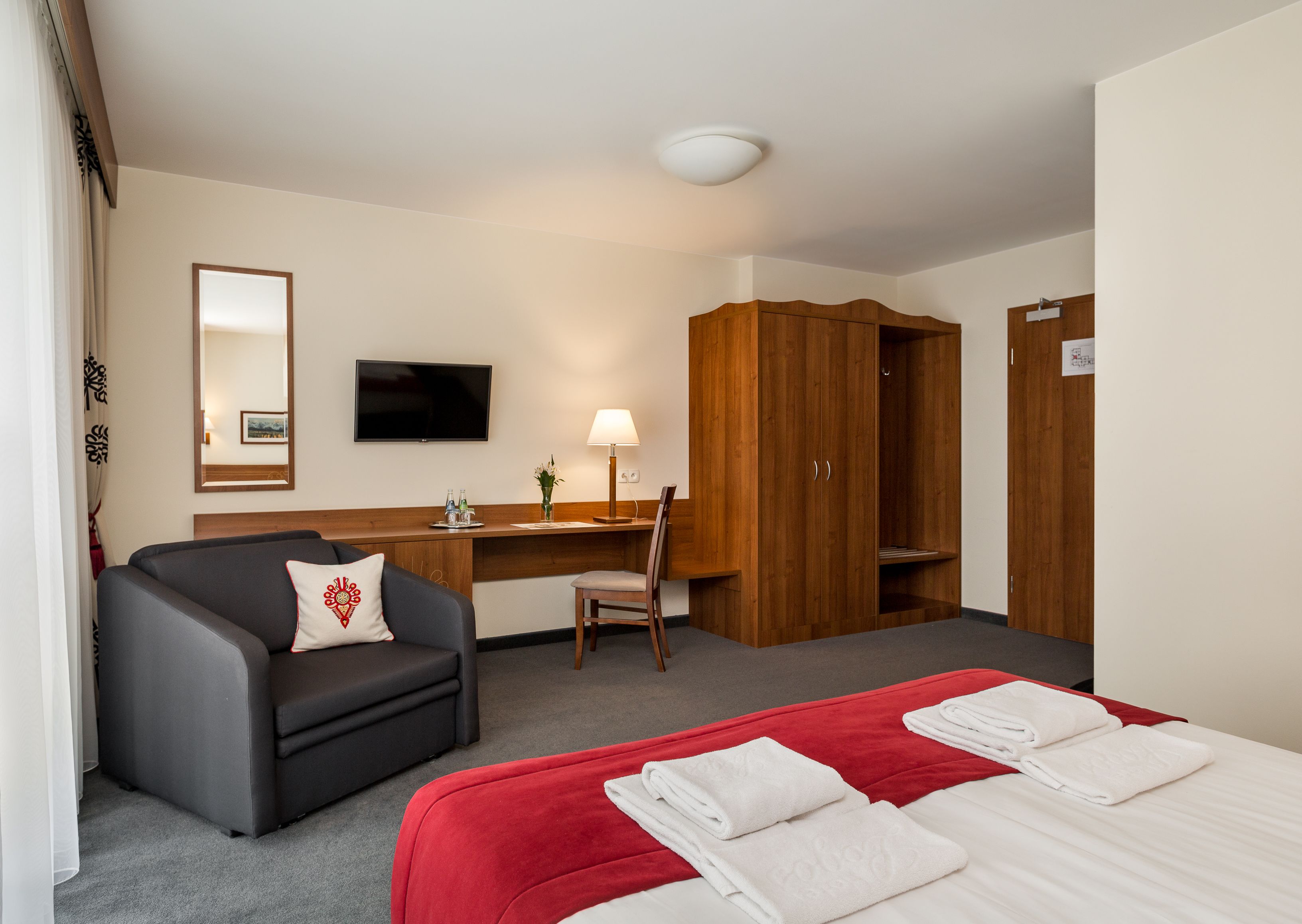 A hotel room with a bed, a desk, a chair, a sofa, a wardrobe, a TV, and a mirror.