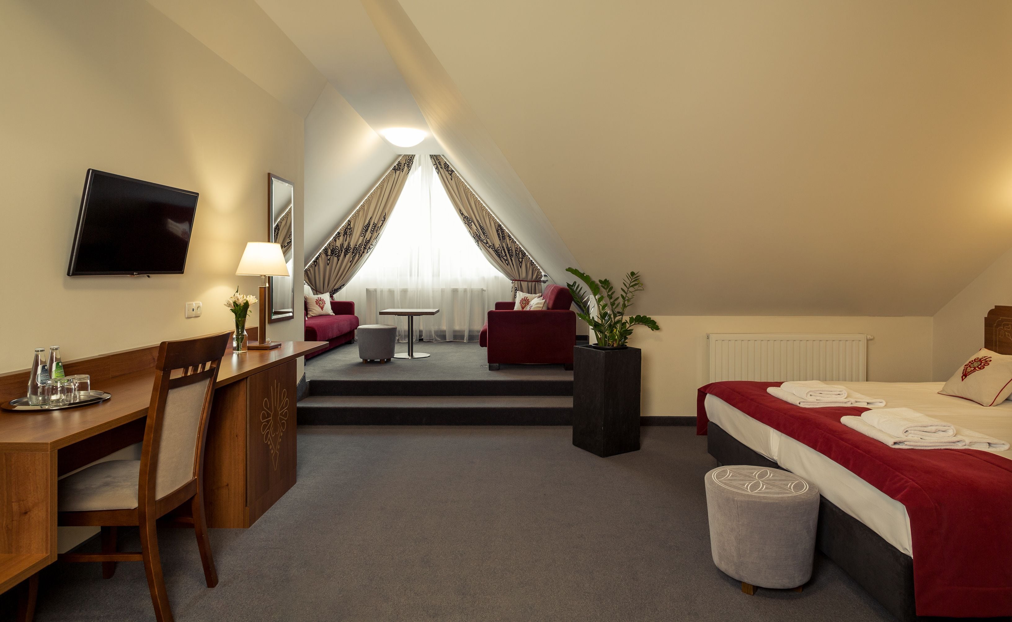 A hotel room with a desk, chair, bed, and a small table with a lamp.