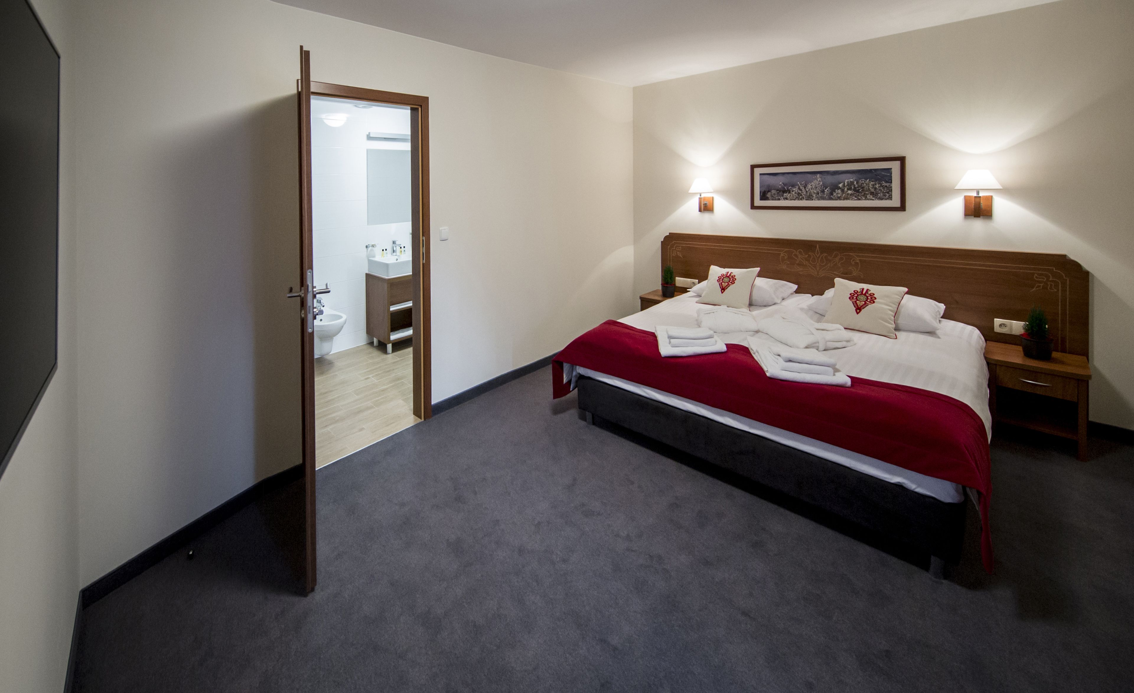 Una habitación de hotel con una cama, una mesita de noche, una lámpara de pared, un cuadro enmarcado y un baño visible a través de una puerta abierta.