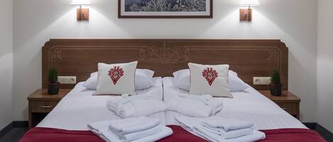 Una cama bien hecha con sábanas blancas y detalles rojos, dos mesitas de noche con lámparas, un cuadro en la pared y una pequeña planta en cada mesita.