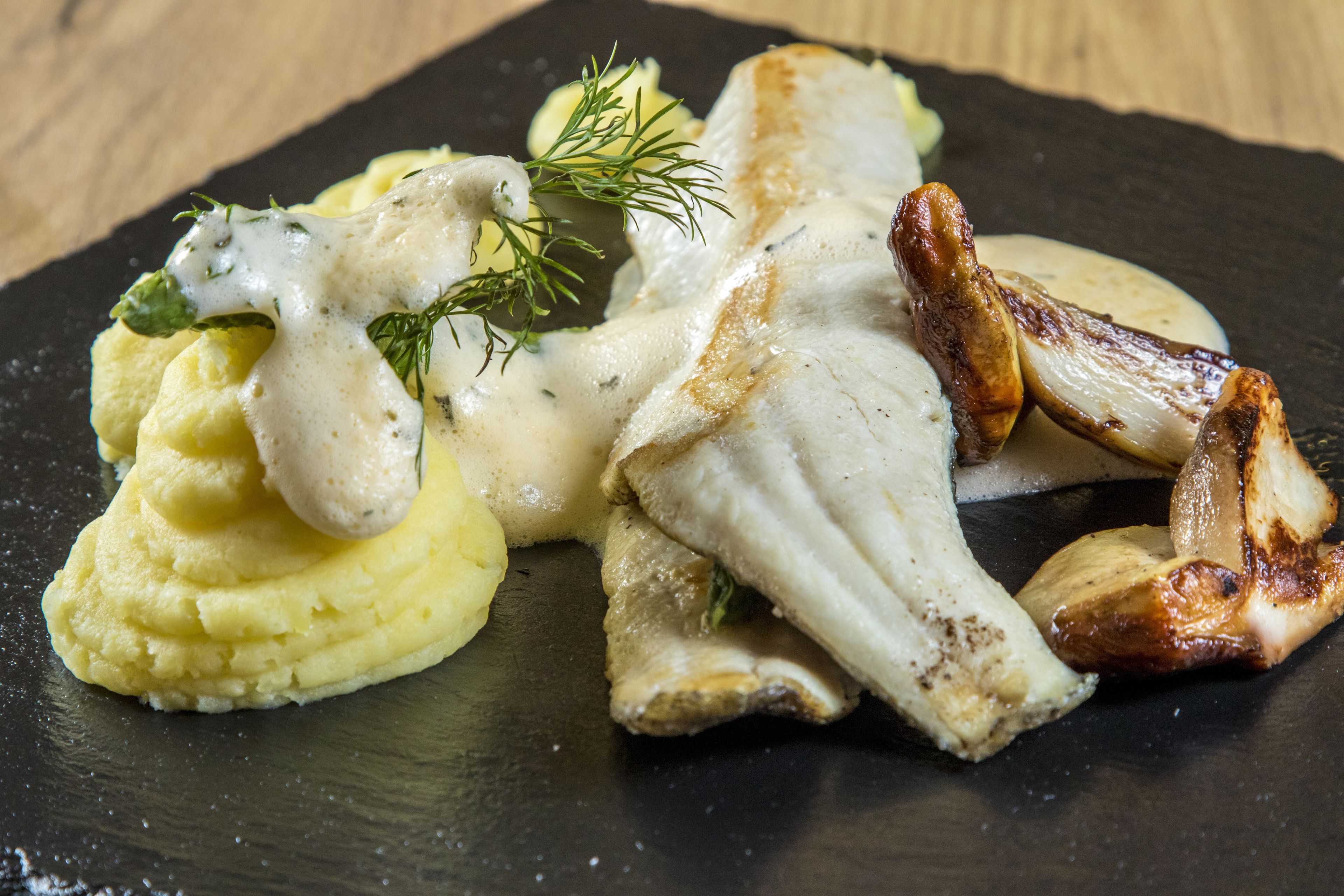 A dish of fish, mashed potatoes, and roasted potatoes on a black plate.