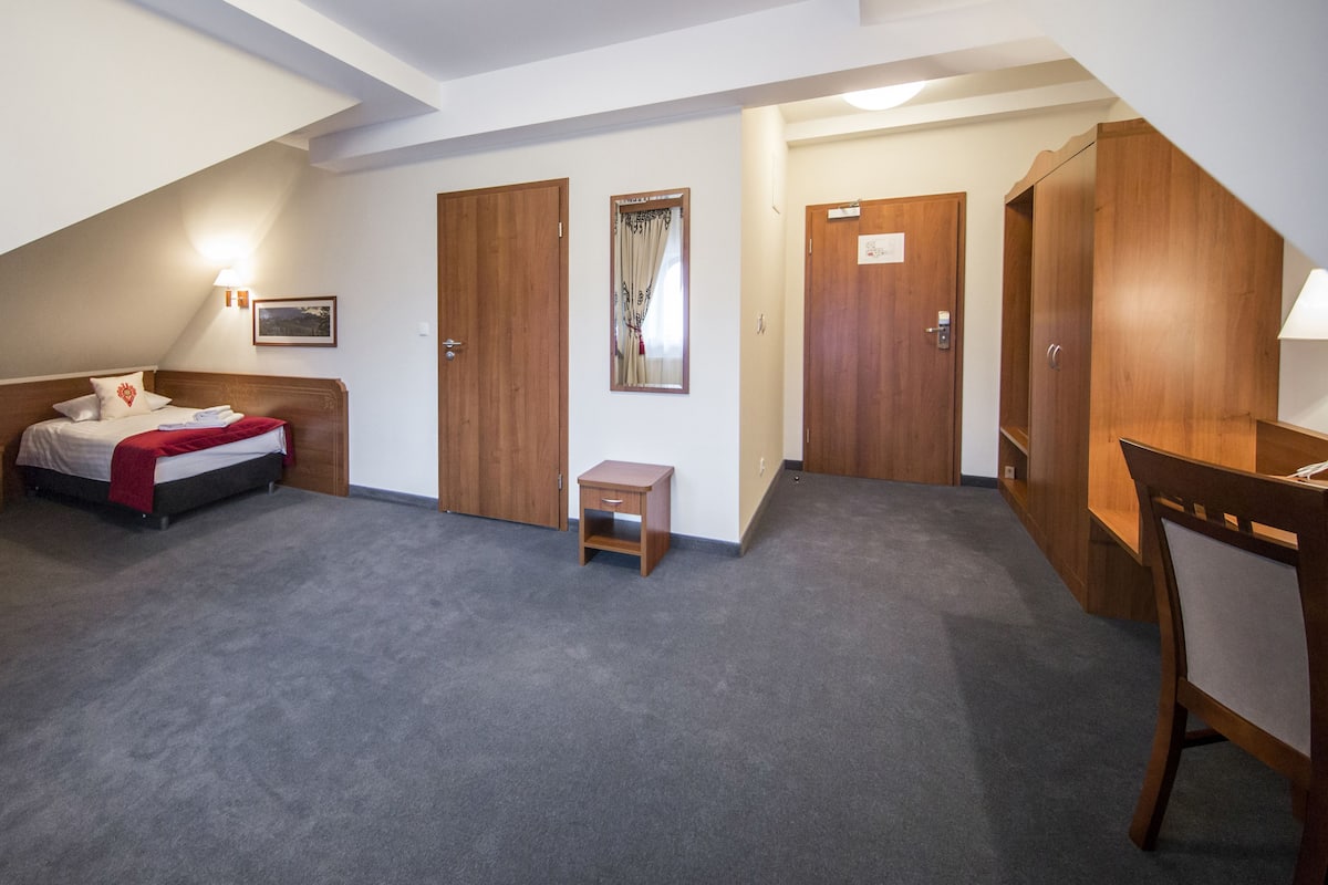 Quarto de hotel com uma cama, uma escrivaninha, uma mesinha e dois guarda-roupas.