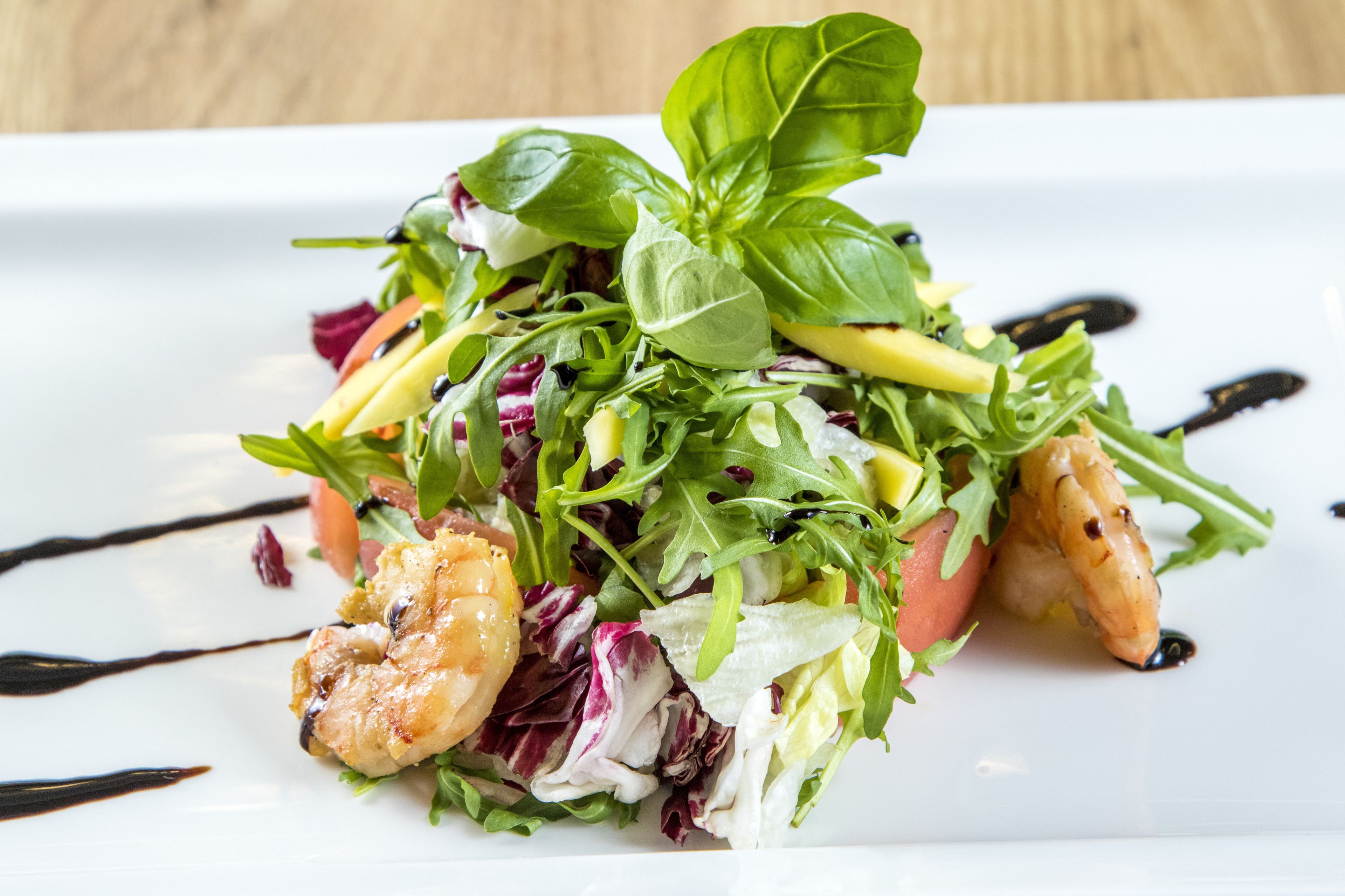 A plate of food with various greens, tomatoes, and shrimp, garnished with basil leaves and drizzled with a dark sauce.