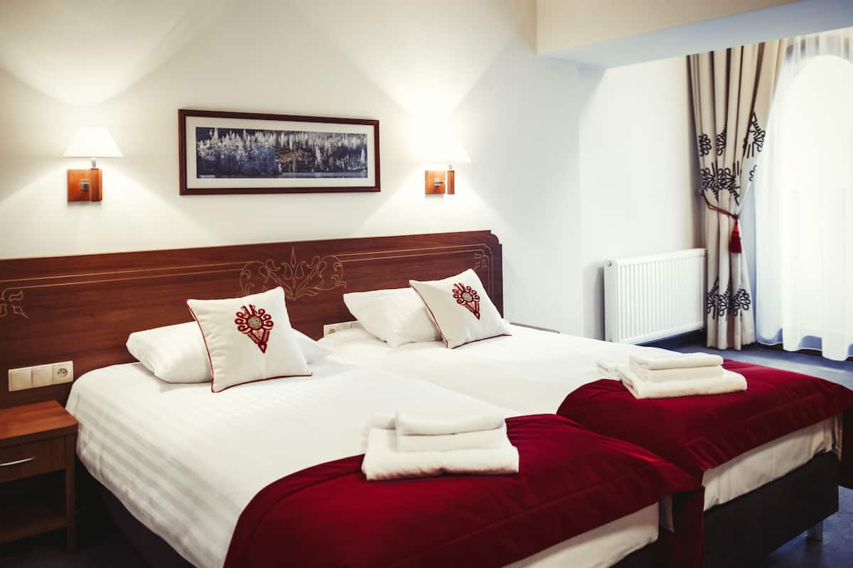 Uma cama de casal com roupa de cama branca e detalhes vermelhos, uma mesinha de cabeceira, um quadro na parede e uma janela com cortinas.
