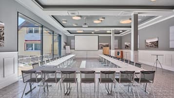 Ett konferensrum med ett långt bord, stolar, en projektorduk och ett stort fönster med utsikt över en byggnad.