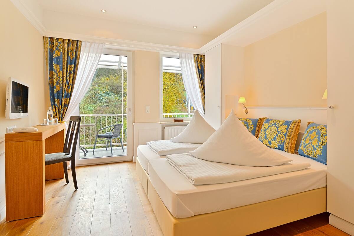 Une chambre à coucher avec un lit, un bureau et une chaise. Il y a un balcon avec vue sur les arbres.
