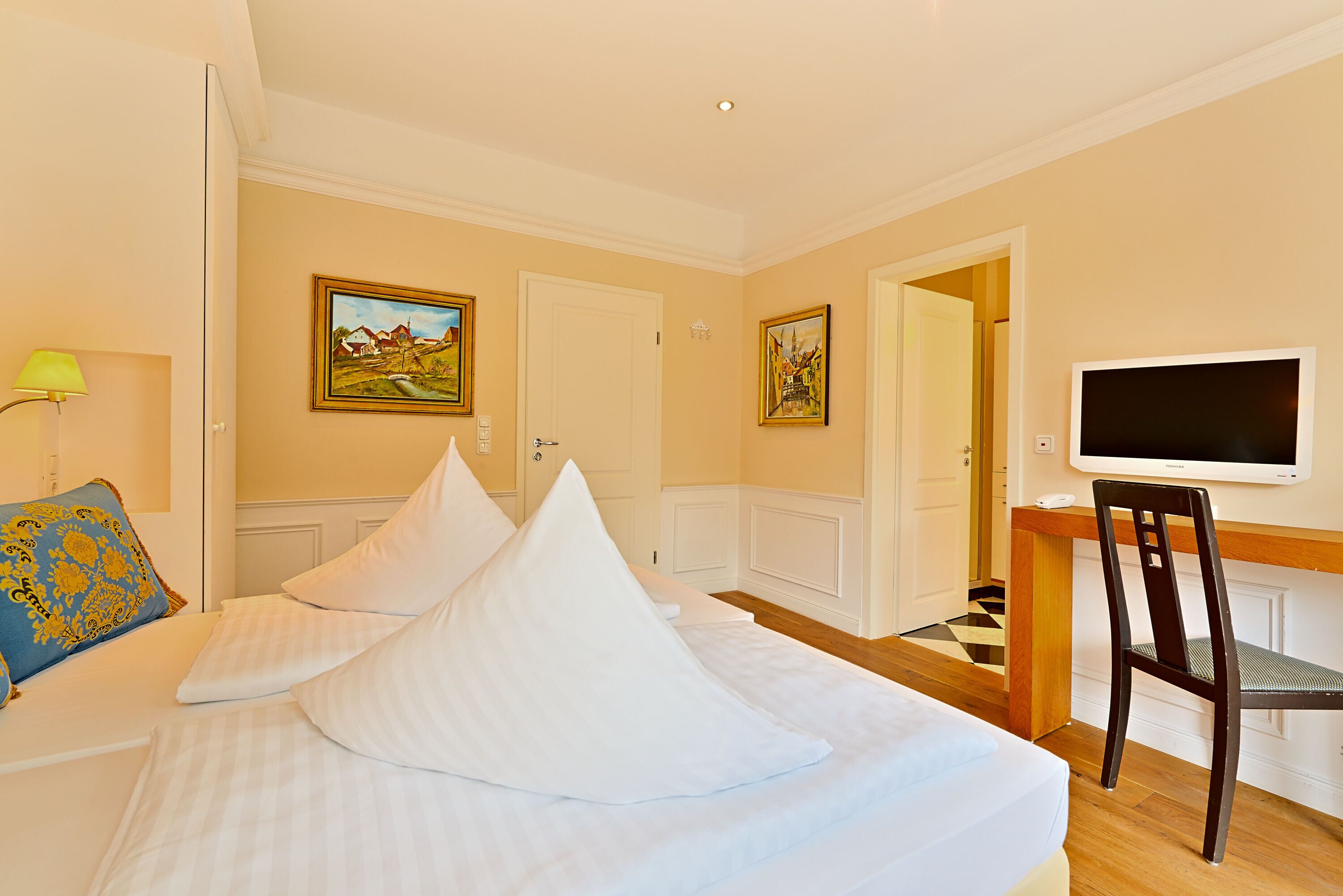 A hotel room with a bed, a desk, a chair, a television, and two paintings on the wall.