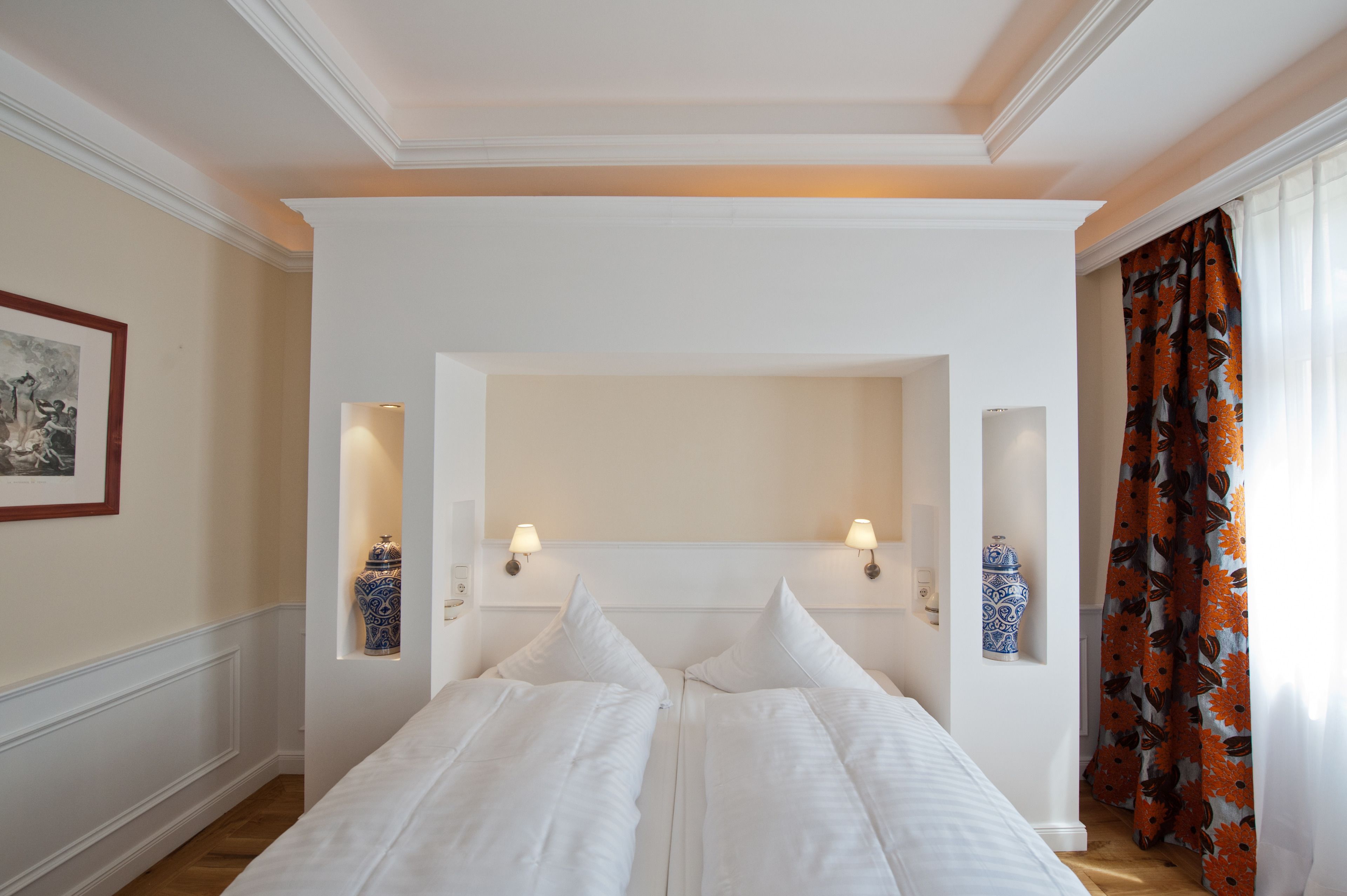 Een hotelkamer met een groot bed, twee wandlampen, een lijst met een afbeelding en een raam met gordijnen.