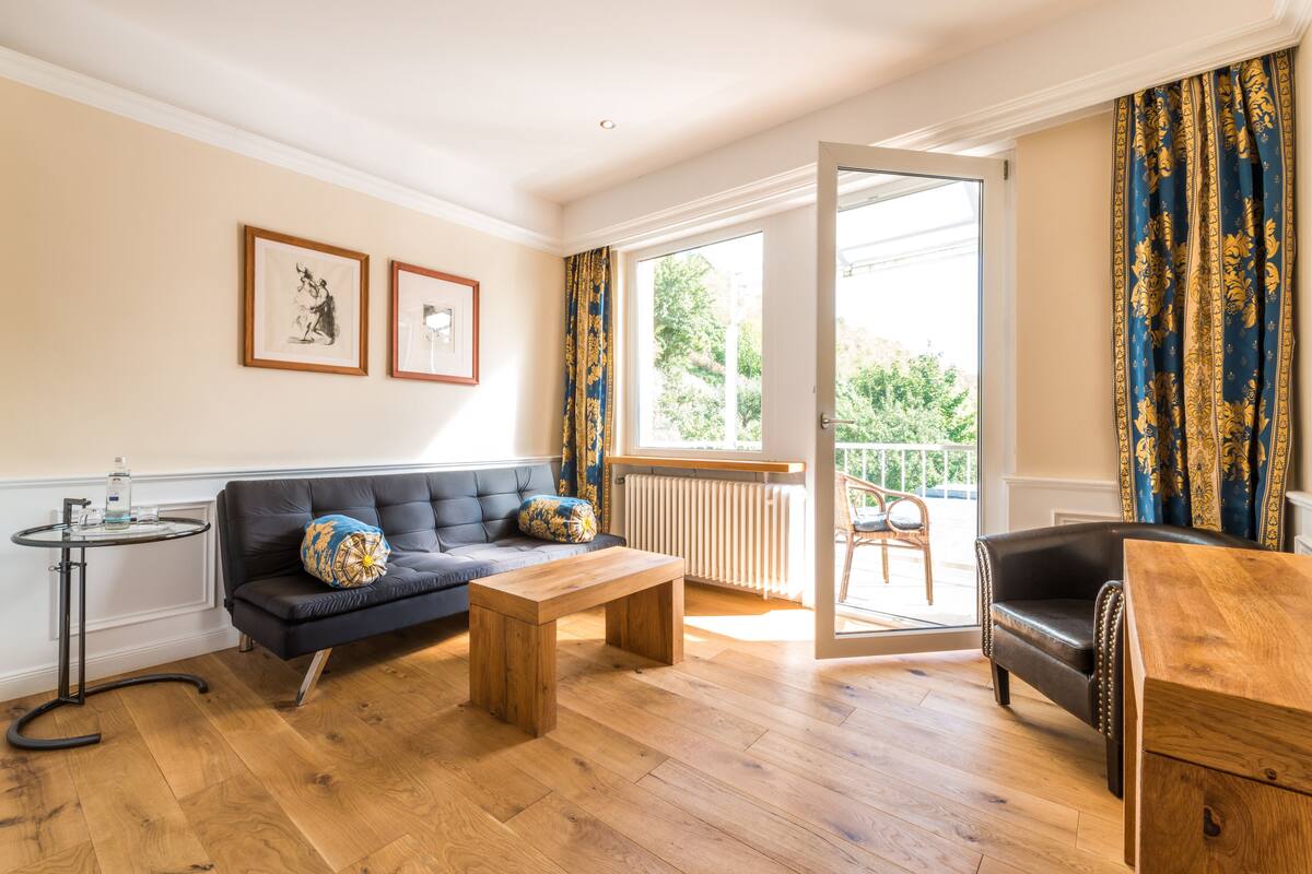 Un salon avec un canapé, une table basse en bois, un fauteuil en cuir noir et une table d’appoint sur laquelle se trouve une bouteille. Deux tableaux encadrés ornent le mur, il y a une fenêtre avec des rideaux et, par la fenêtre, on aperçoit un paysage verdoyant.