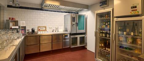 A kitchen with a coffee machine, a refrigerator stocked with beverages, and a counter with glasses and condiments.