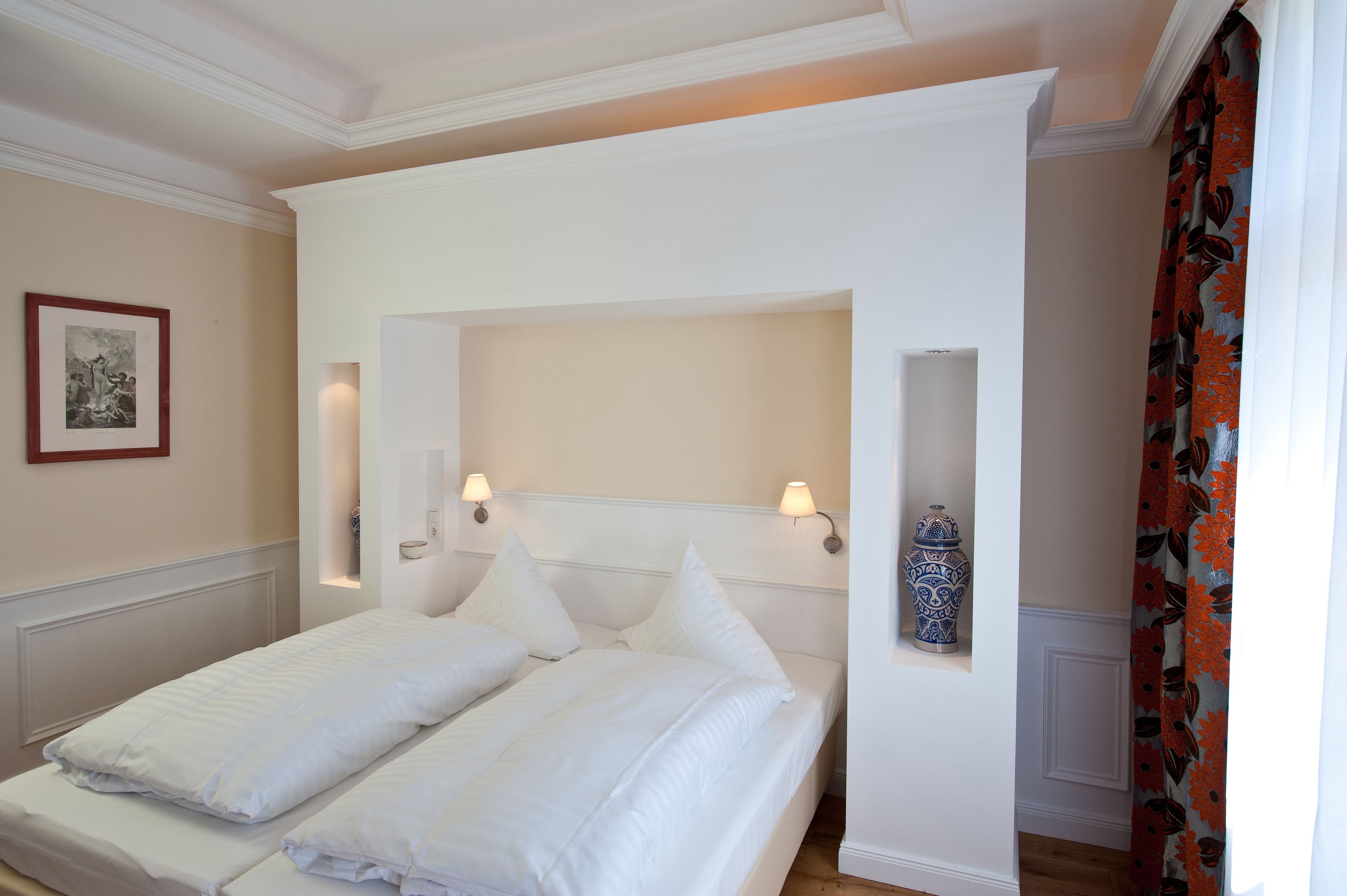 Una cama doble con sábanas blancas, dos lámparas de pared y un jarrón decorativo en una estantería.