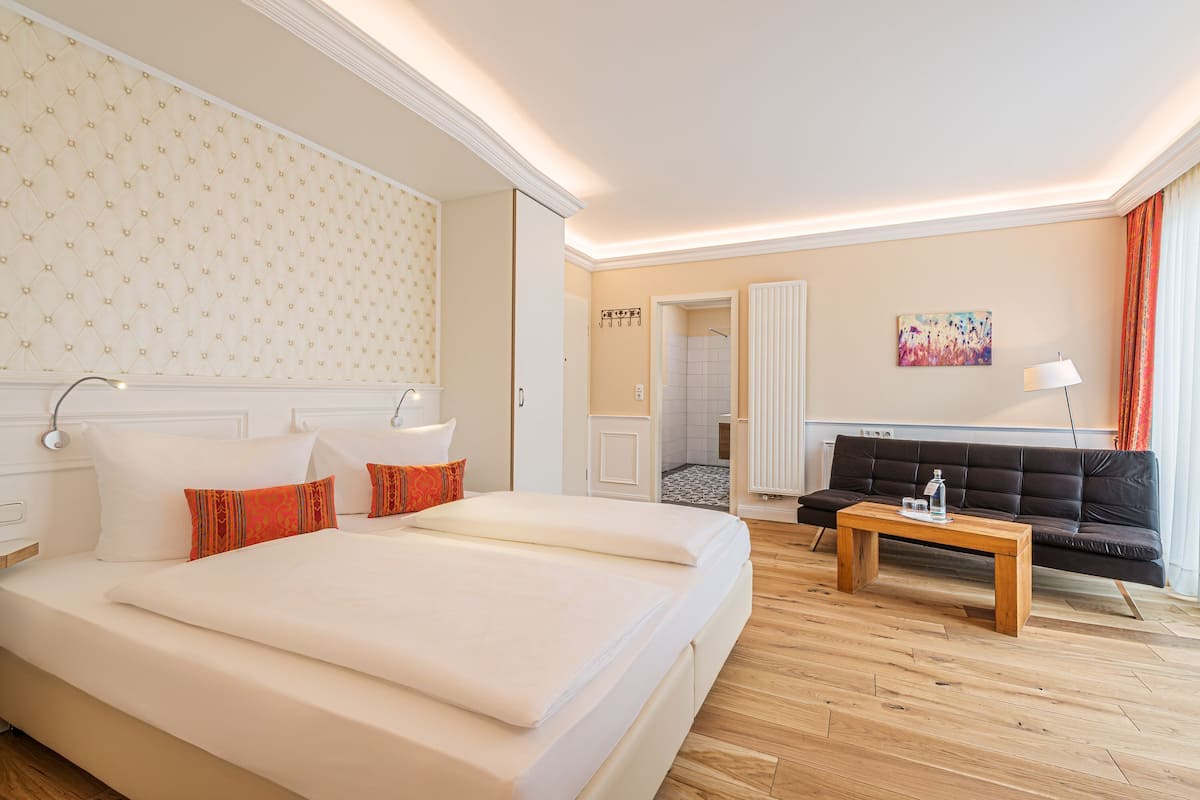 Une chambre d’hôtel avec un grand lit, une table basse en bois, un canapé noir et une salle de bain visible à travers une porte ouverte.