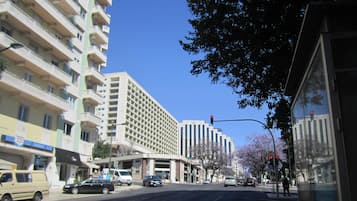 Ein mehrstöckiges Gebäude mit einer rasterförmigen Fassade, eine Bushaltestelle rechts und Fahrzeuge auf der Straße.