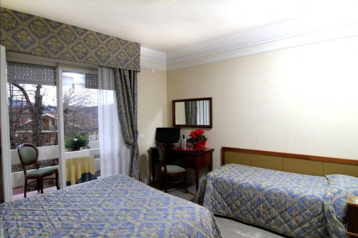 Habitación de hotel con dos camas, un escritorio con computadora, una silla, una ventana con cortinas y un espejo en la pared.