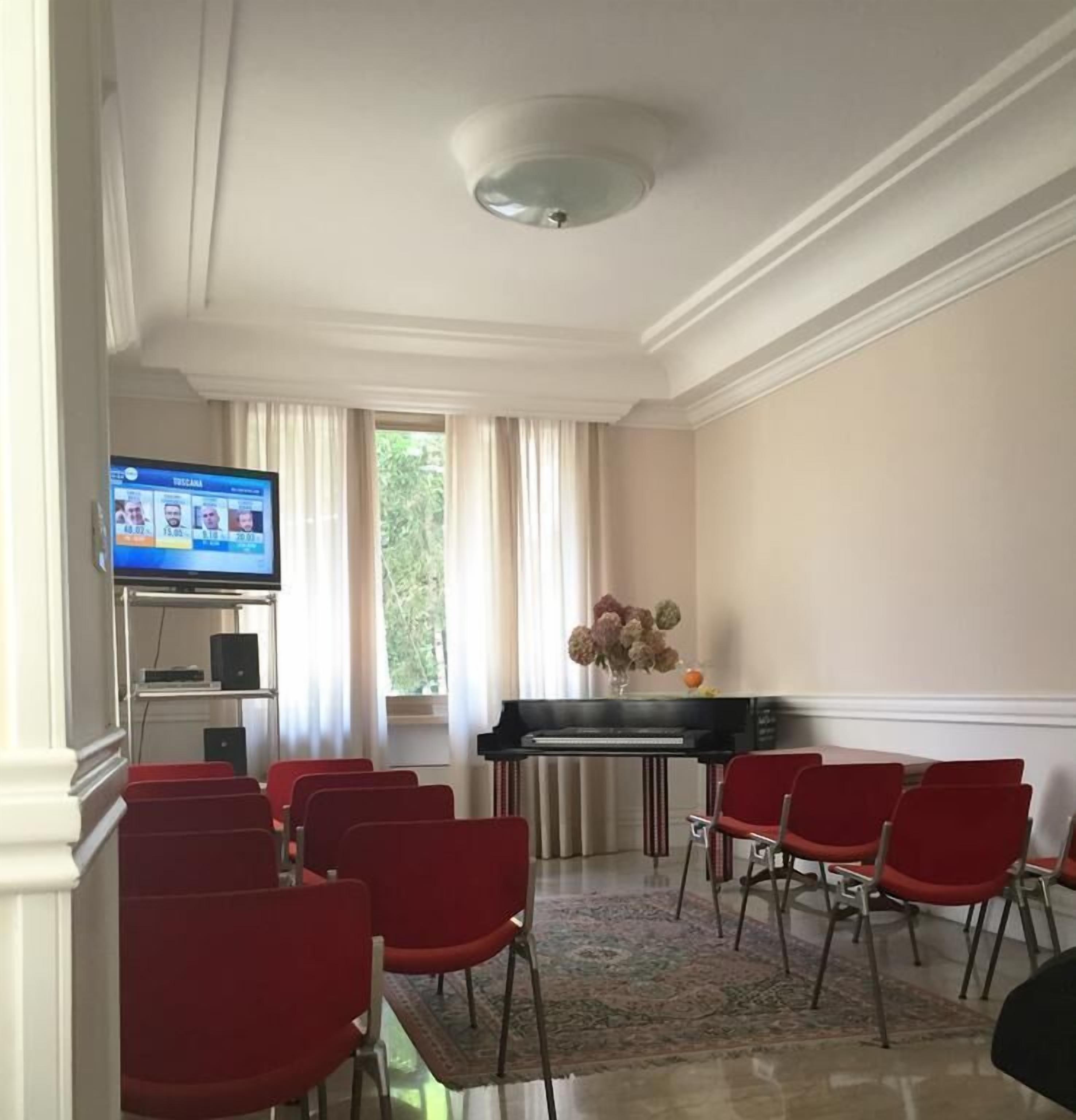 A conference room with a piano, red chairs, a television displaying a sports event, and a window with curtains.