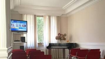 A conference room with a piano, red chairs, a television displaying a sports event, and a window with curtains.
