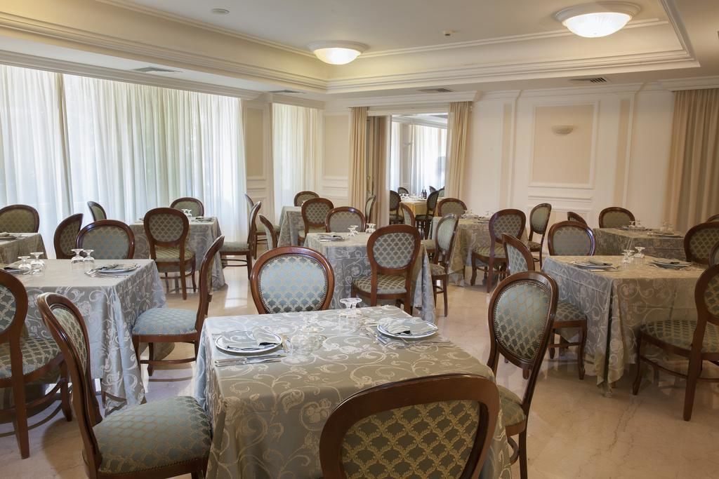 A formal dining room with multiple tables and chairs, arranged in a symmetrical layout, with large windows and curtains.