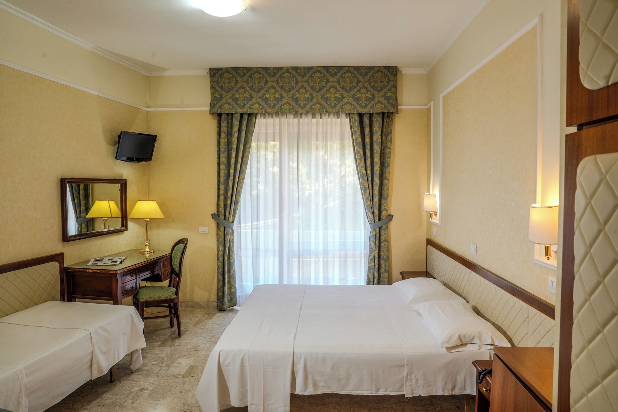 Une chambre d’hôtel avec deux lits, un bureau avec une chaise, une télévision et une fenêtre avec des rideaux.