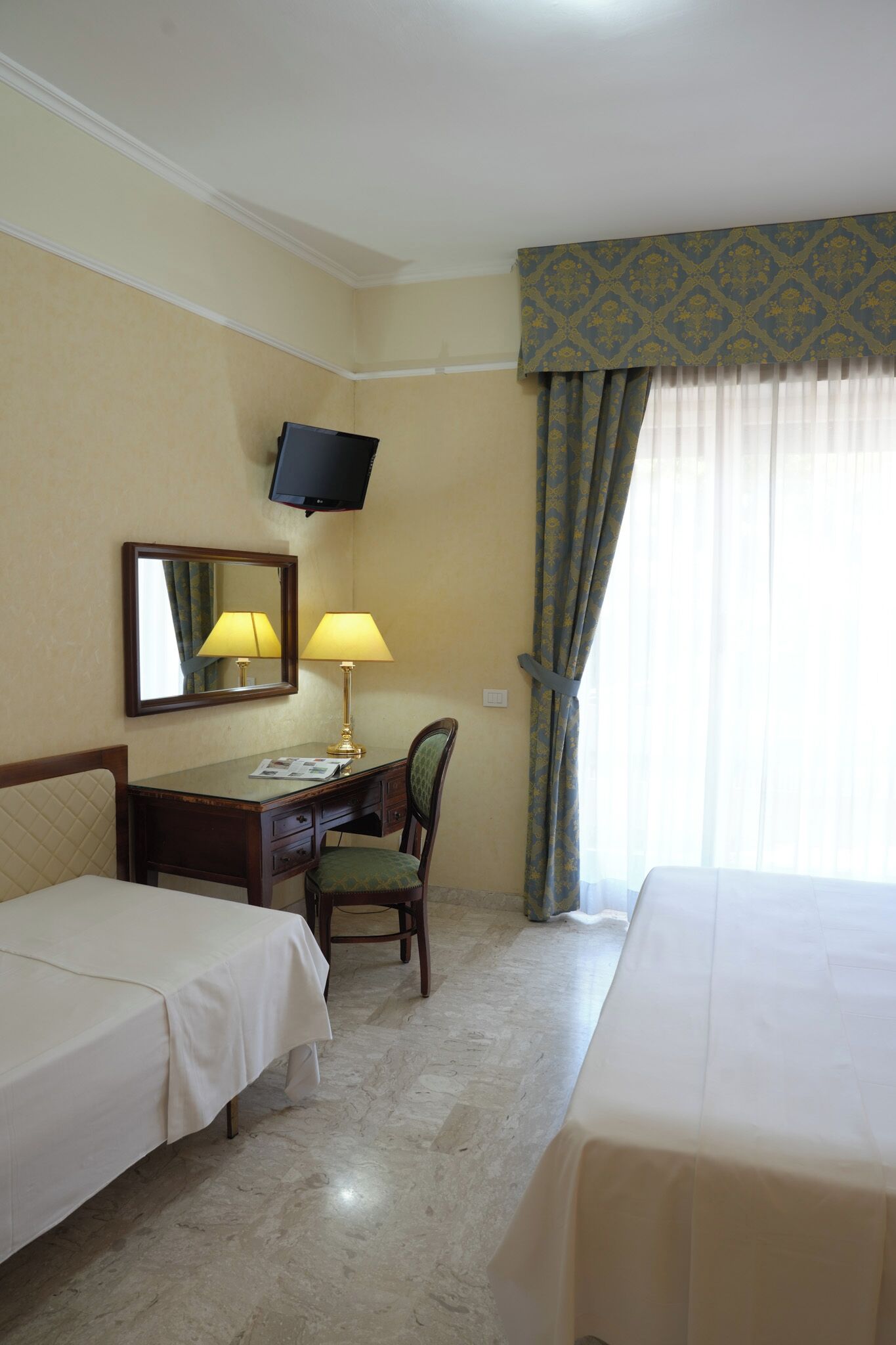 Ein Hotelzimmer mit zwei Betten, einem Schreibtisch mit Stuhl, einem an der Wand befestigten Fernseher und einem Fenster mit Vorhängen.