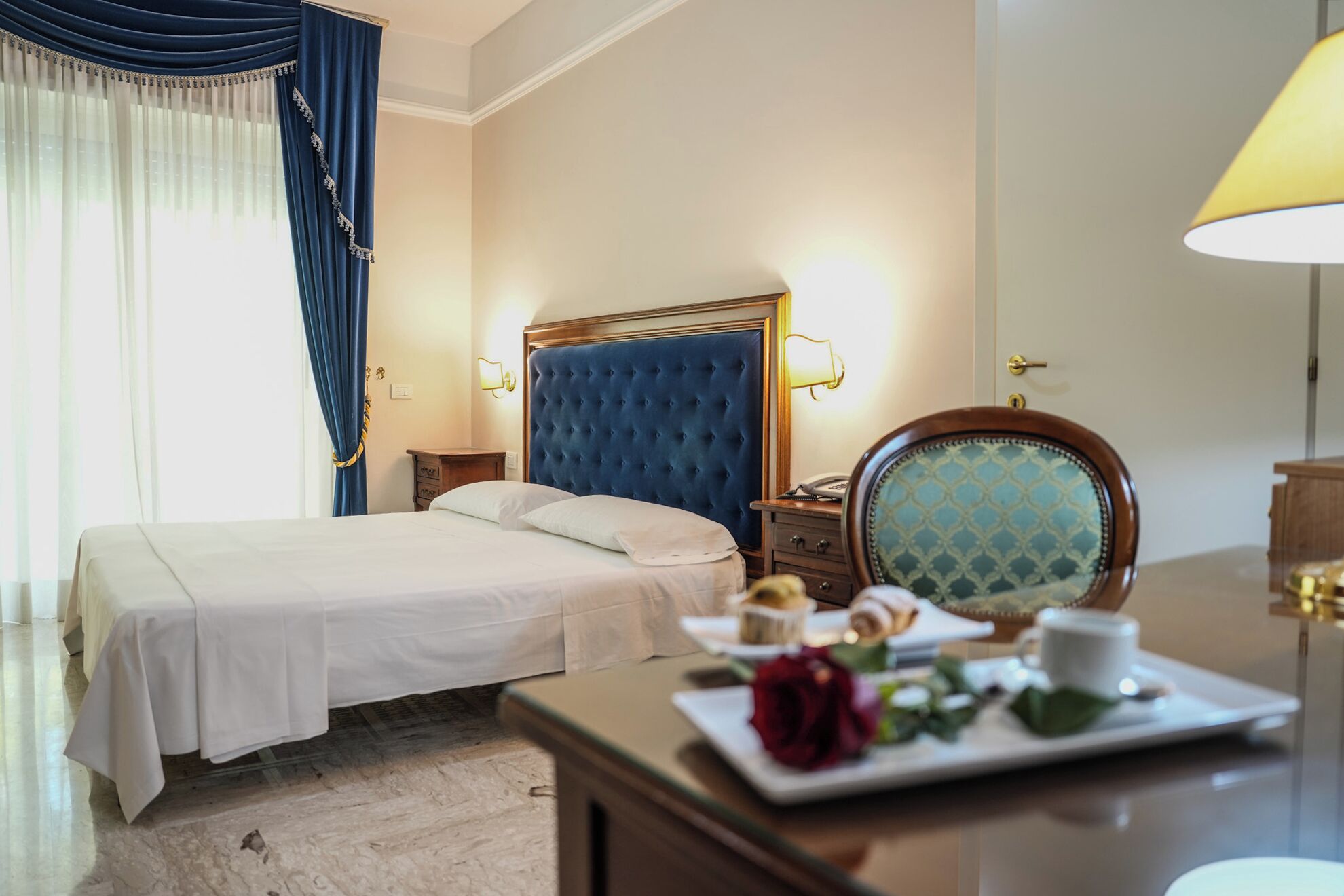 A hotel room with a bed, bedside table, lamp, and a tray with a rose and pastries.