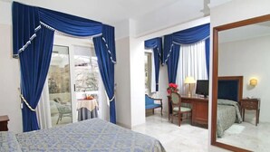 Habitación de hotel con cama, escritorio, silla, espejo y balcón con cortinas azules.