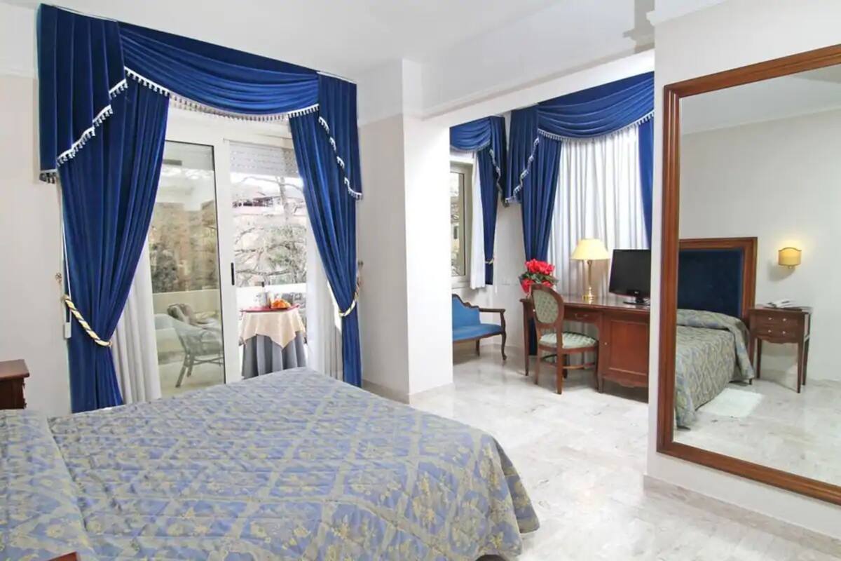 Habitación de hotel con cama, escritorio, silla, espejo y balcón con cortinas azules.