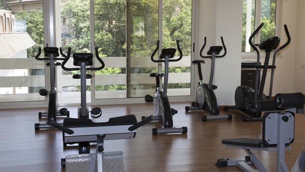 Un gimnasio con diferentes equipos de ejercicio, ventanas amplias y vistas al exterior con vegetación.