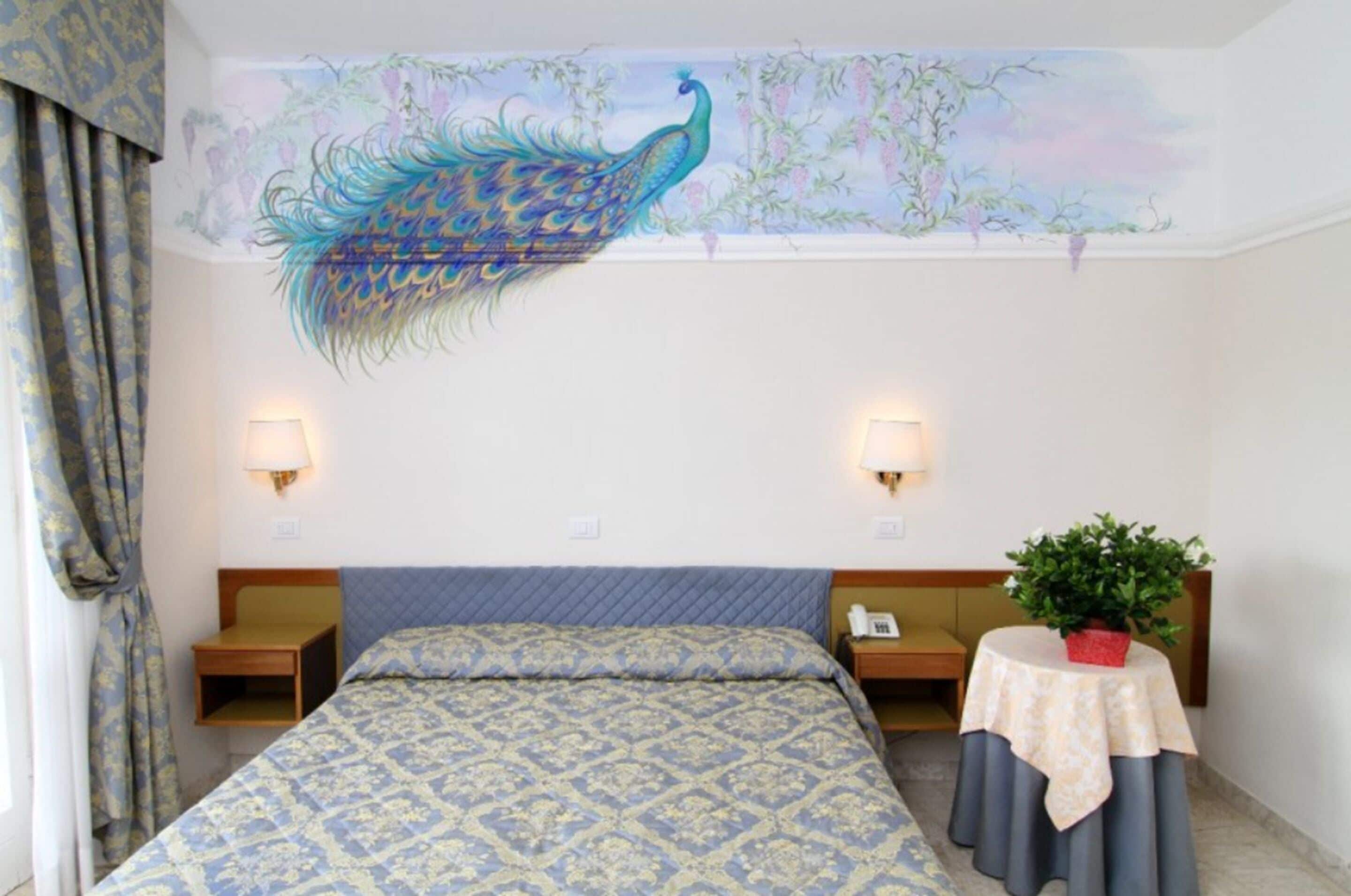 A hotel room with a large peacock mural on the ceiling, a bed with patterned bedding, bedside lamps, a nightstand, a small table with a plant, and a telephone.