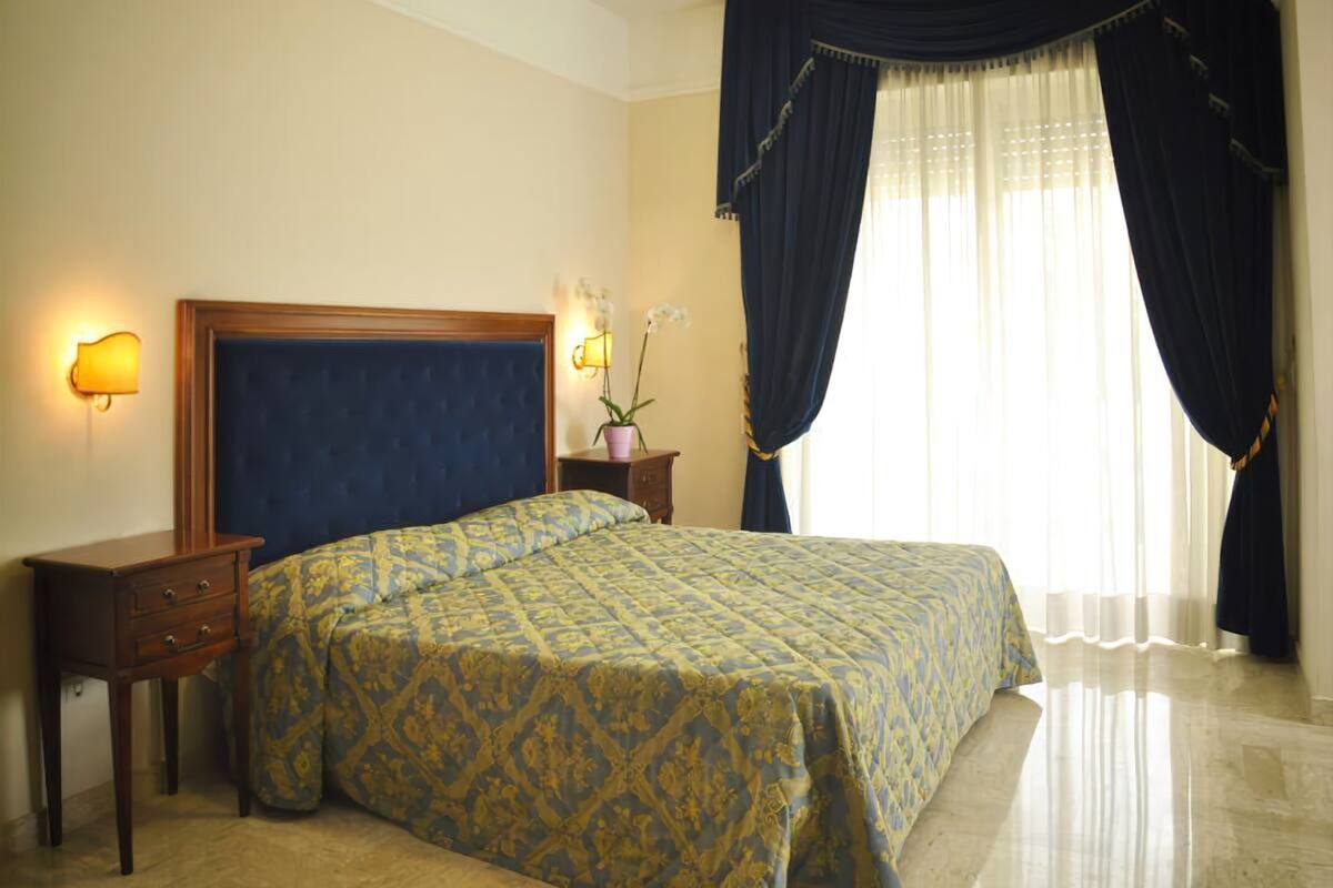 Habitación de hotel con una cama grande, una mesita de noche, una lámpara y una ventana con cortinas.