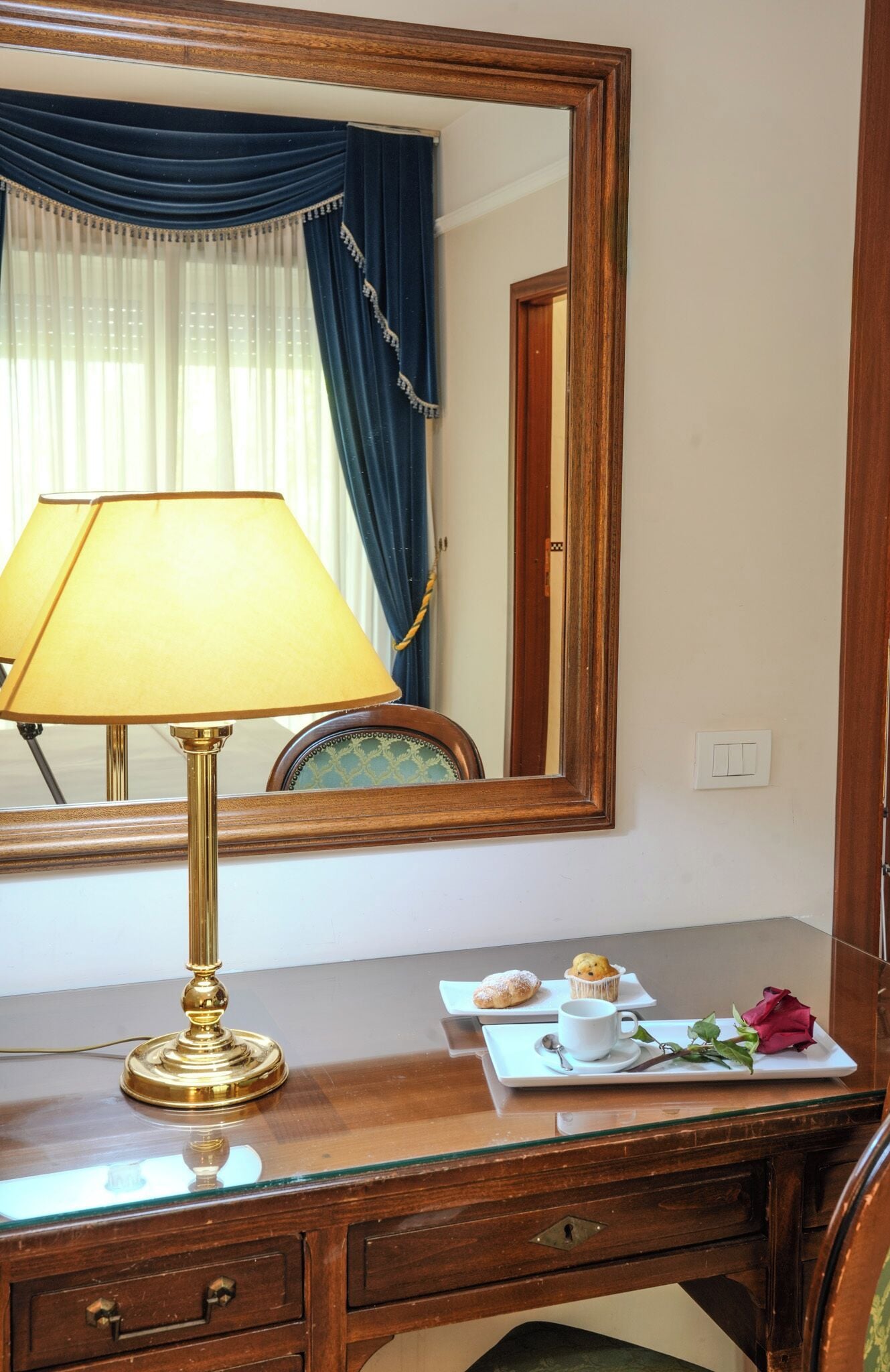 A wooden desk with a brass lamp, a mirror, and a plate with pastries and a rose.