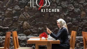 A person seated at a table with a stone wall background and a logo for 'ILOS KITCHEN'. - Hotel Ilos (Bandung)