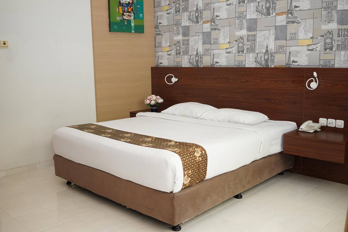 Una cama bien hecha con una colcha estampada, una mesita de noche con un teléfono y una pared con papel tapiz de paisaje urbano.