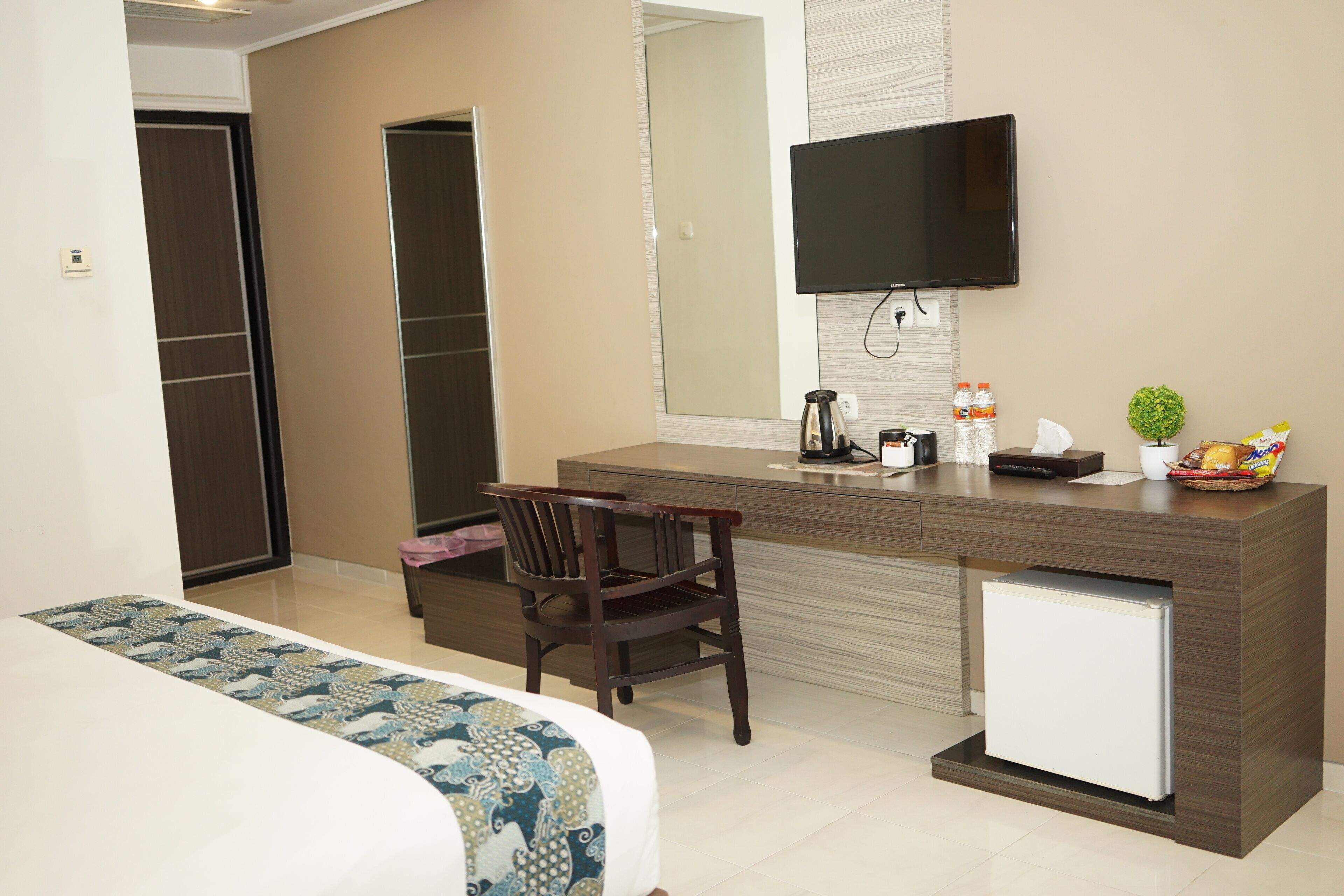 A hotel room with a bed, a desk with a chair, a small refrigerator, a television mounted on the wall, and a mirror.