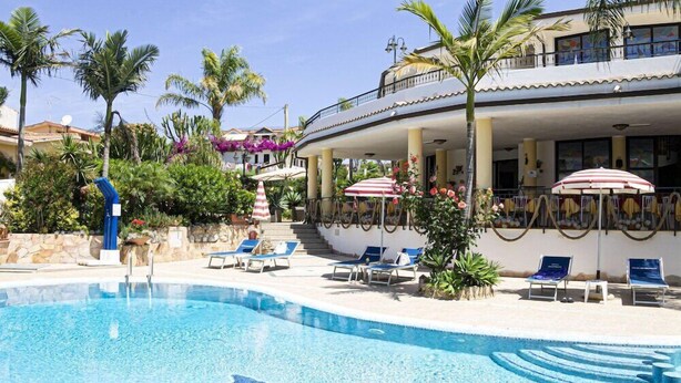 A pool area with lounge chairs and umbrellas, surrounded by palm trees and a building with balconies. - Hotel Incoronato (Capo Vaticano)