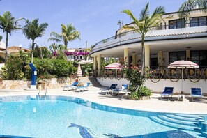 A pool area with lounge chairs and umbrellas, surrounded by palm trees and a building with balconies.