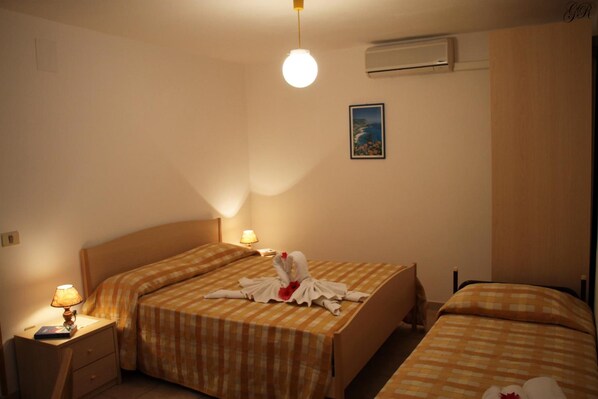 A hotel room with two beds, each with a checkered bedspread, a bedside table with a lamp, and a wall-mounted air conditioner.