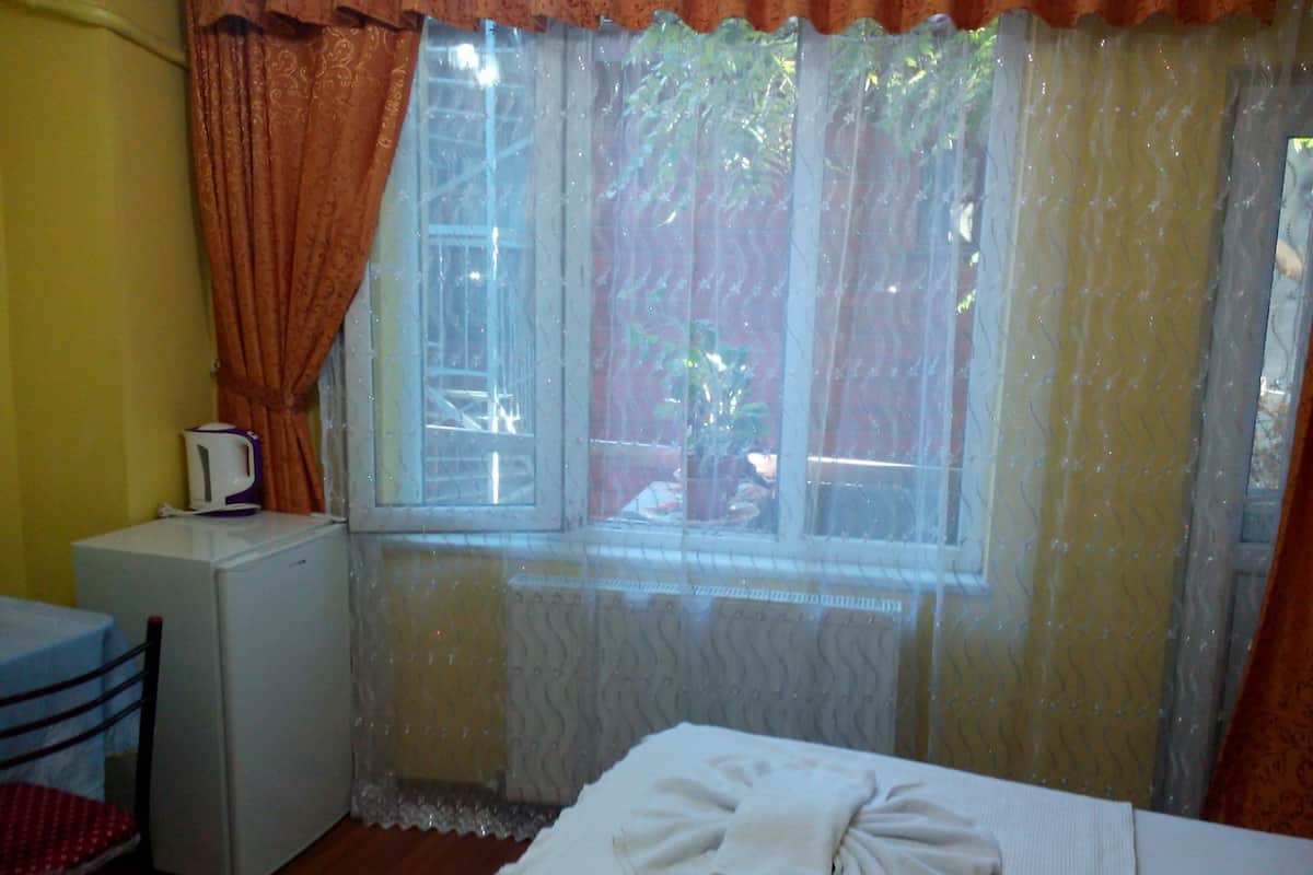 A small room with a bed, a table, a chair, a kettle, and a window with curtains.