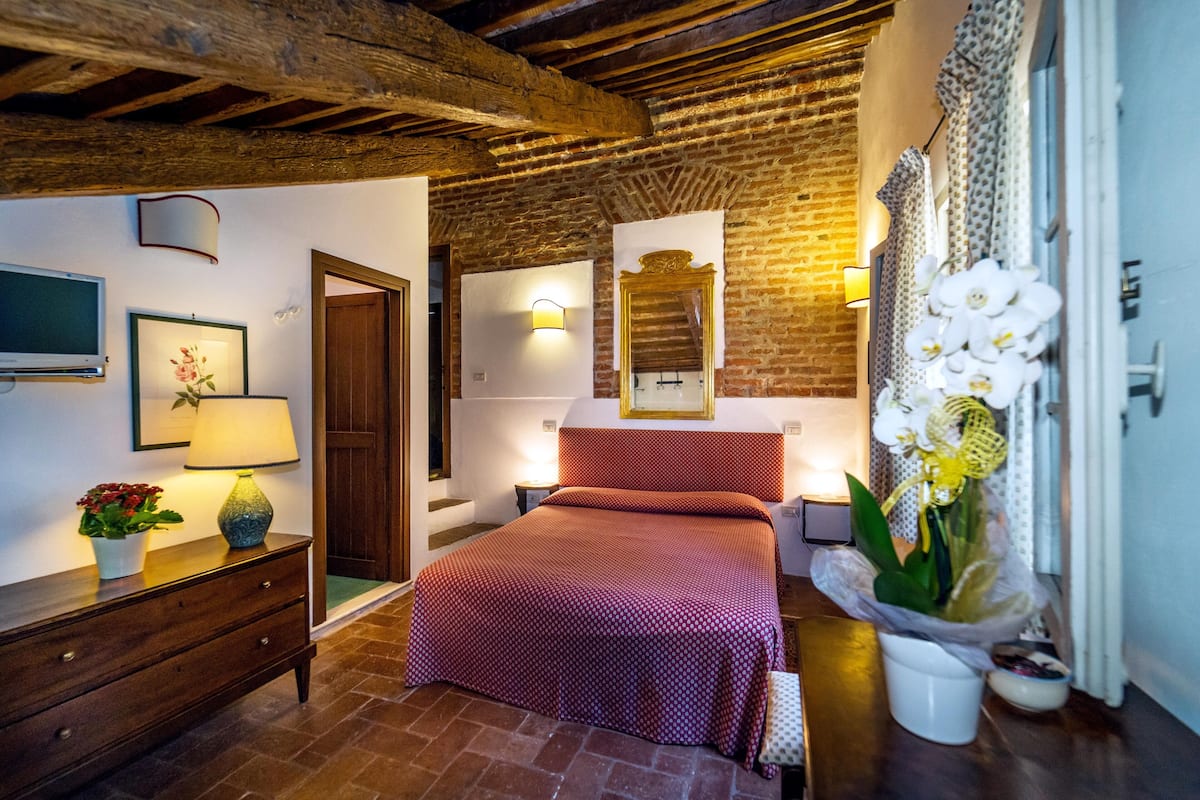Une chambre avec un mur en briques, des poutres en bois, un lit recouvert d’une couverture rouge, une commode, un miroir et un pot d’orchidée.