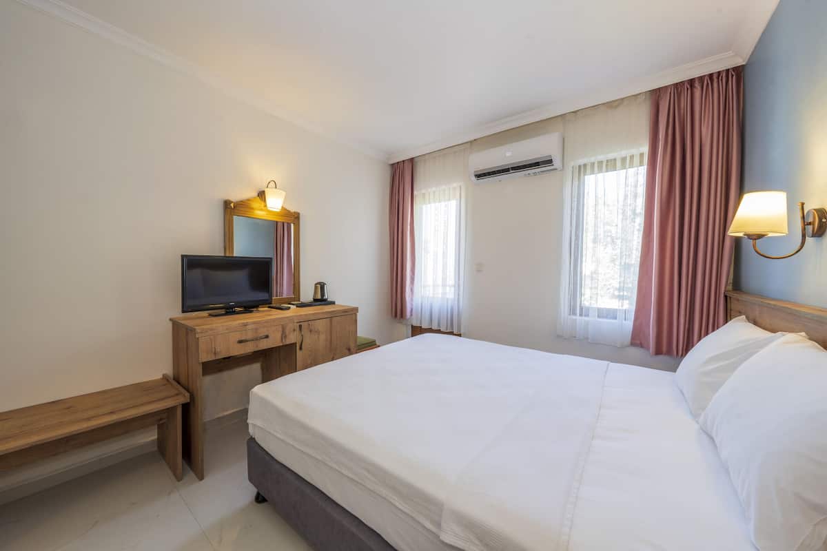 A hotel room with a bed, a wooden desk with a television, a mirror, and a wall-mounted air conditioner.
