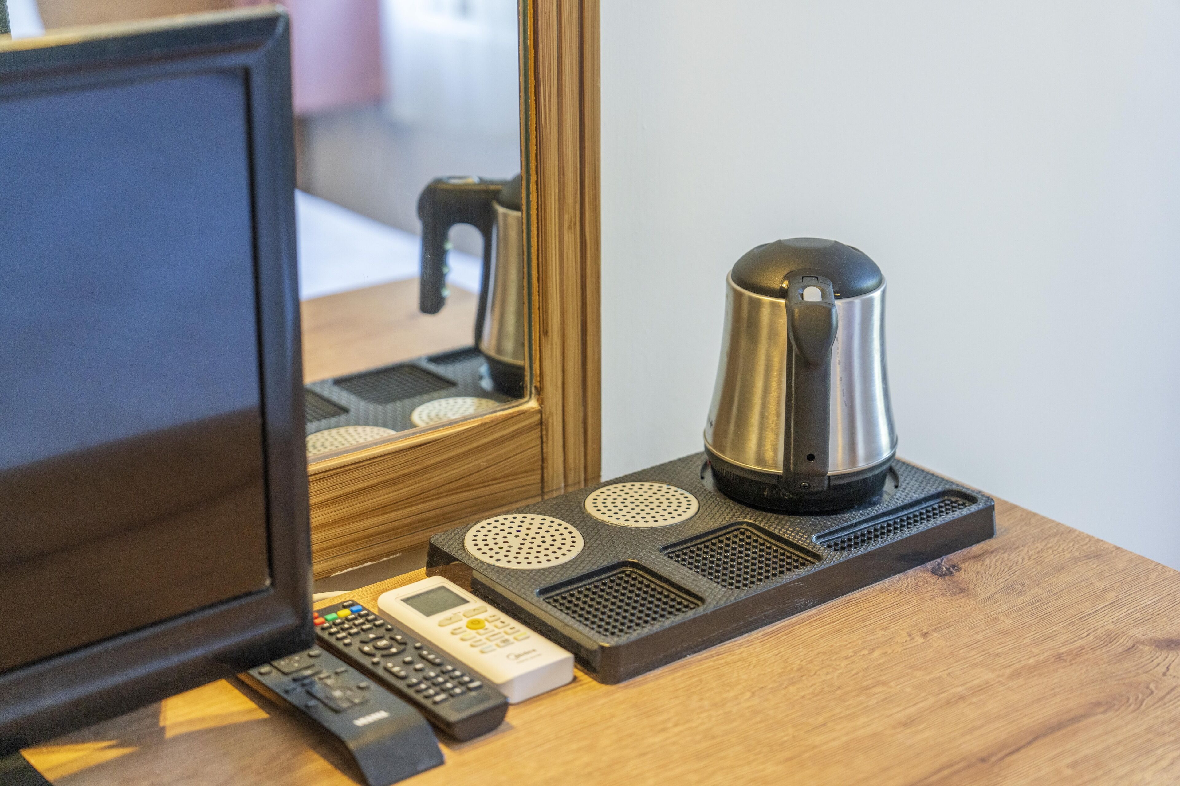 A hotel room with a wooden desk, a coffee maker, a telephone, and a mirror reflecting a bed and a chair.