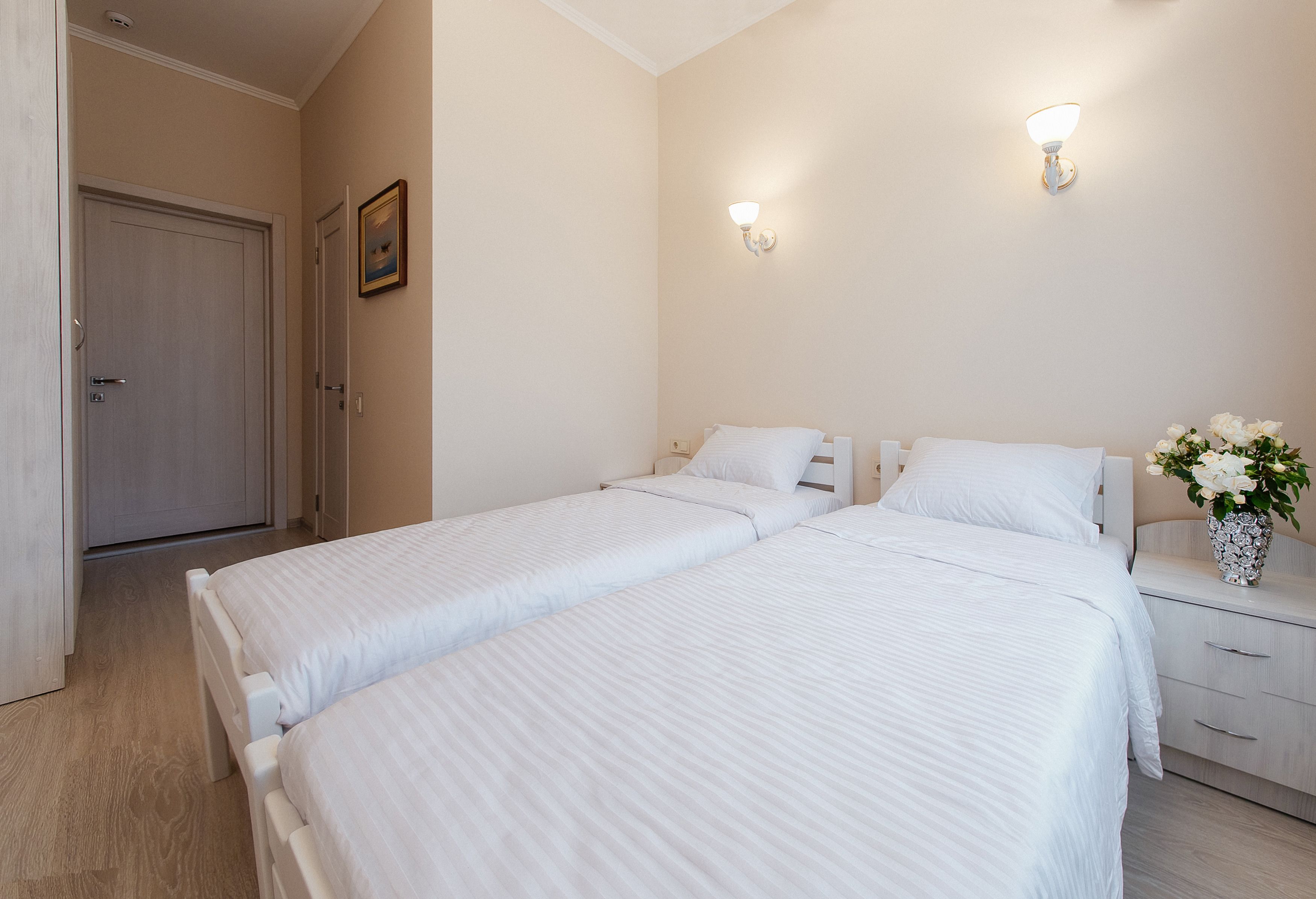 Une chambre d’hôtel avec deux lits simples, une table de chevet, une commode, un vase avec des fleurs et une porte donnant sur une autre pièce.