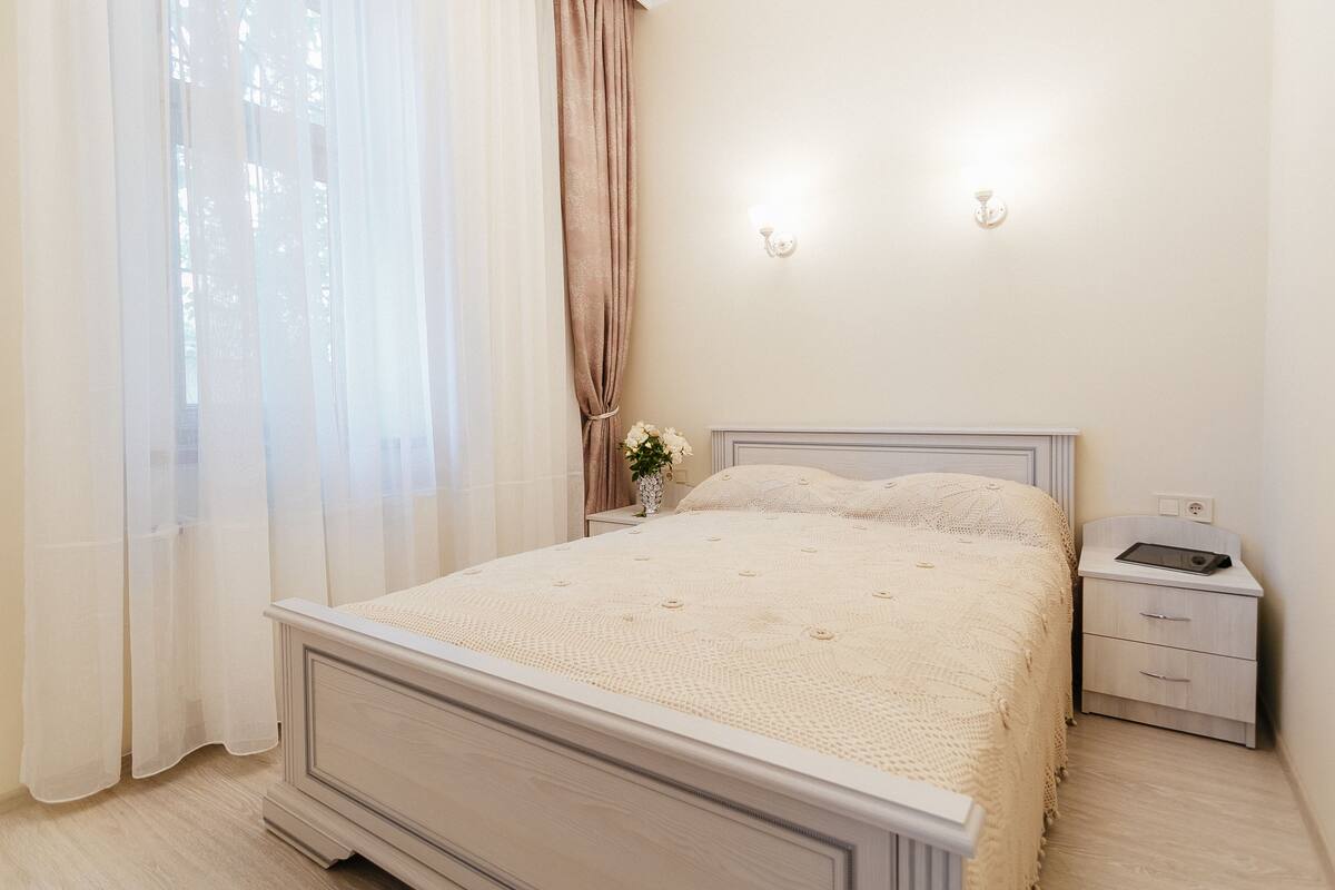 A bedroom with a bed, a nightstand, a lamp, and a window with sheer curtains.