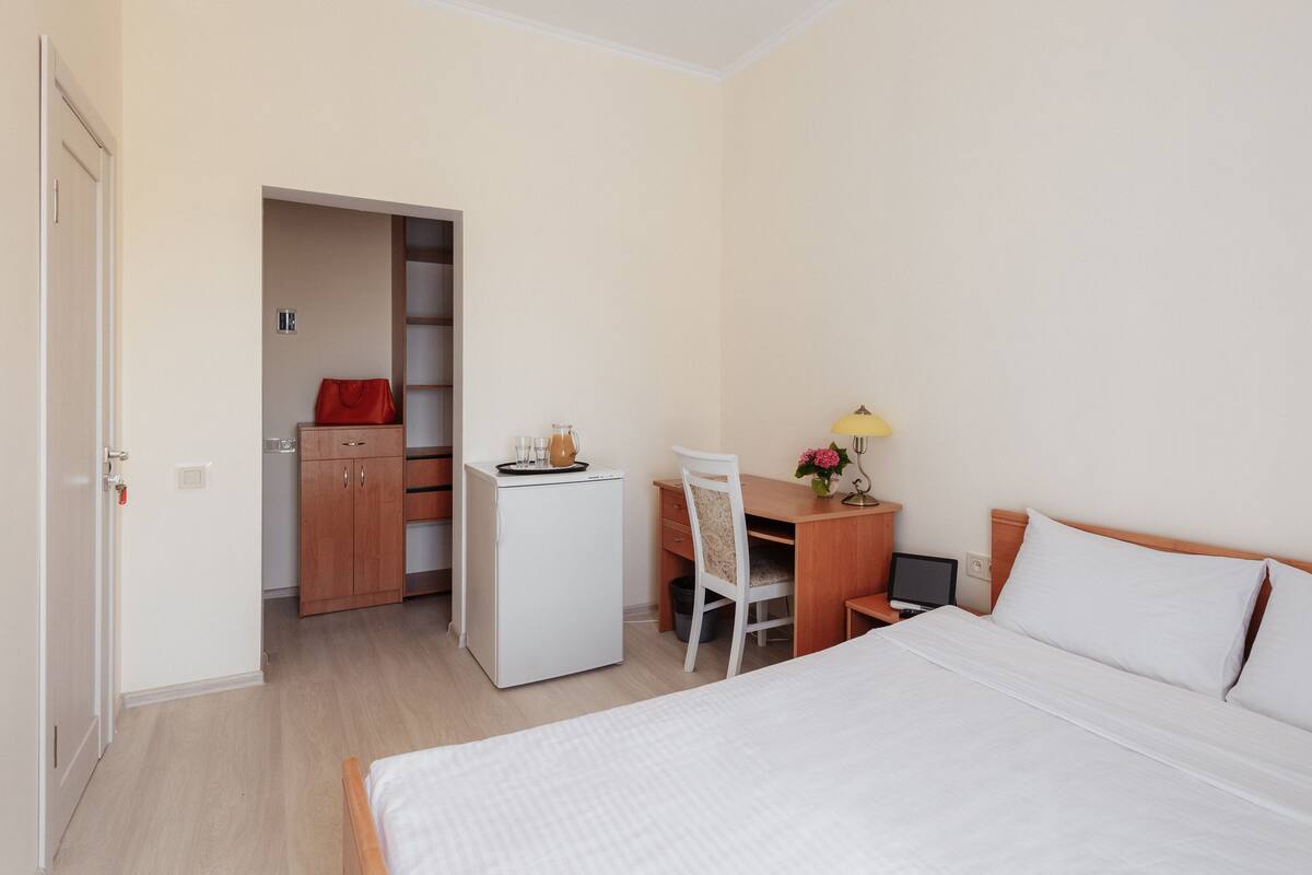 A hotel room with a bed, a desk with a chair, a small refrigerator, and a wardrobe.
