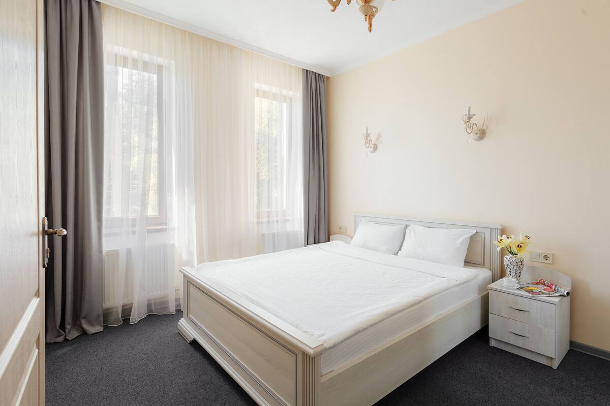 A neatly made bed with white linens, a bedside table with a vase of flowers, and a window with sheer curtains.