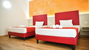 Quarto de hotel com duas camas, cada uma com cabeceira vermelha e roupa de cama branca. O quarto possui piso de madeira e uma parede de destaque laranja.