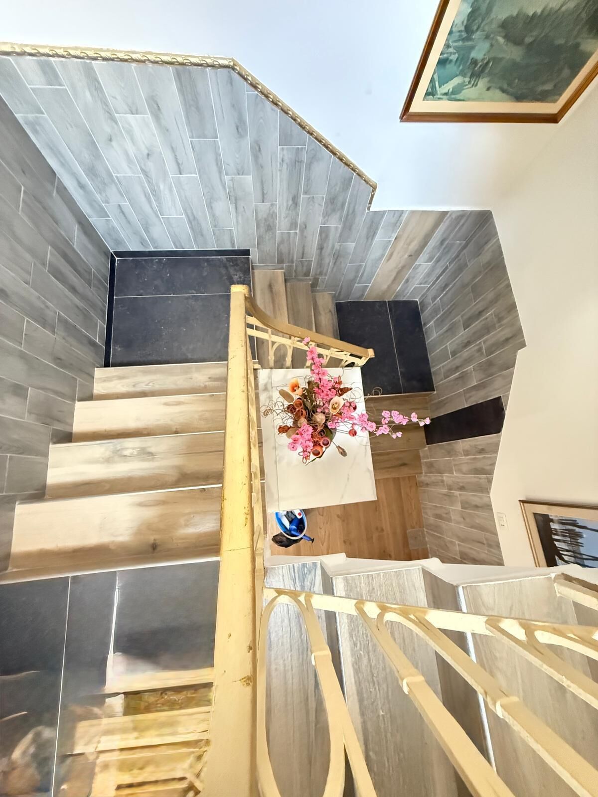 A staircase with wooden steps and a metal railing, leading to a landing with a floral arrangement and a framed picture on the wall.