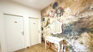 Quarto com piso de madeira, duas portas brancas, toucador branco com espelho e parede texturizada com desenhos florais e abstratos.
