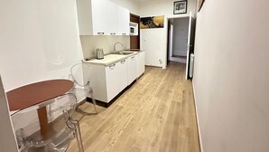 Cozinha compacta com armários brancos, micro-ondas e pia. Piso de madeira e pequena mesa de jantar com cadeiras.