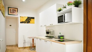 Cozinha compacta com armários brancos, micro-ondas e bancada pequena. Há uma porta que dá para outro cômodo e uma cadeira perto da pia.
